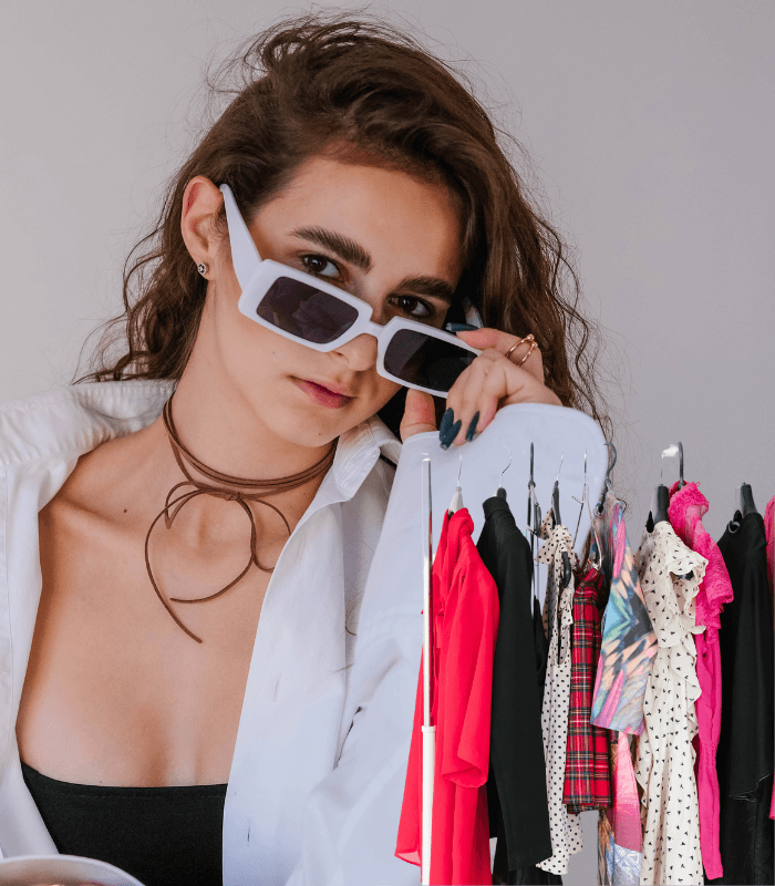 A young woman with long brown hair and sunglasses poses confidently in front of a clothing rack filled with various colorful outfits.