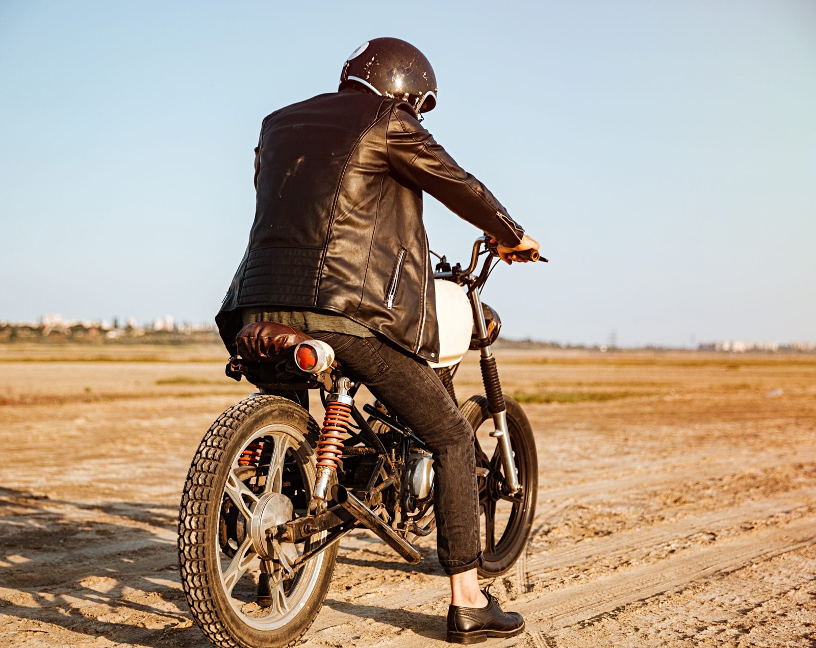 Young man in a black jacket and glasses riding a retro motorcycle, seen from the back.