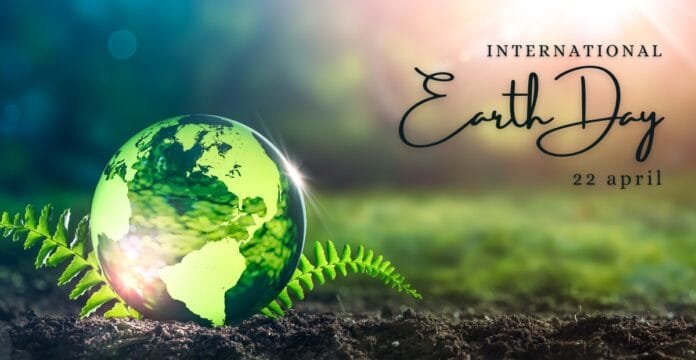 A stylized green globe with continents is nestled among ferns, symbolizing Earth Day. The background is softly blurred with a warm light and natural landscape.