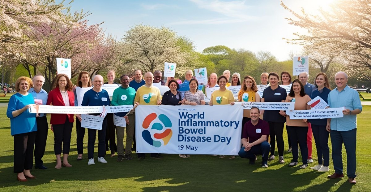 World Inflammatory Bowel Disease Day – 19 May A diverse group of people gathered outdoors in a park with blossoming trees, holding a banner and signs promoting World Inflammatory Bowel Disease Day on 19 May.