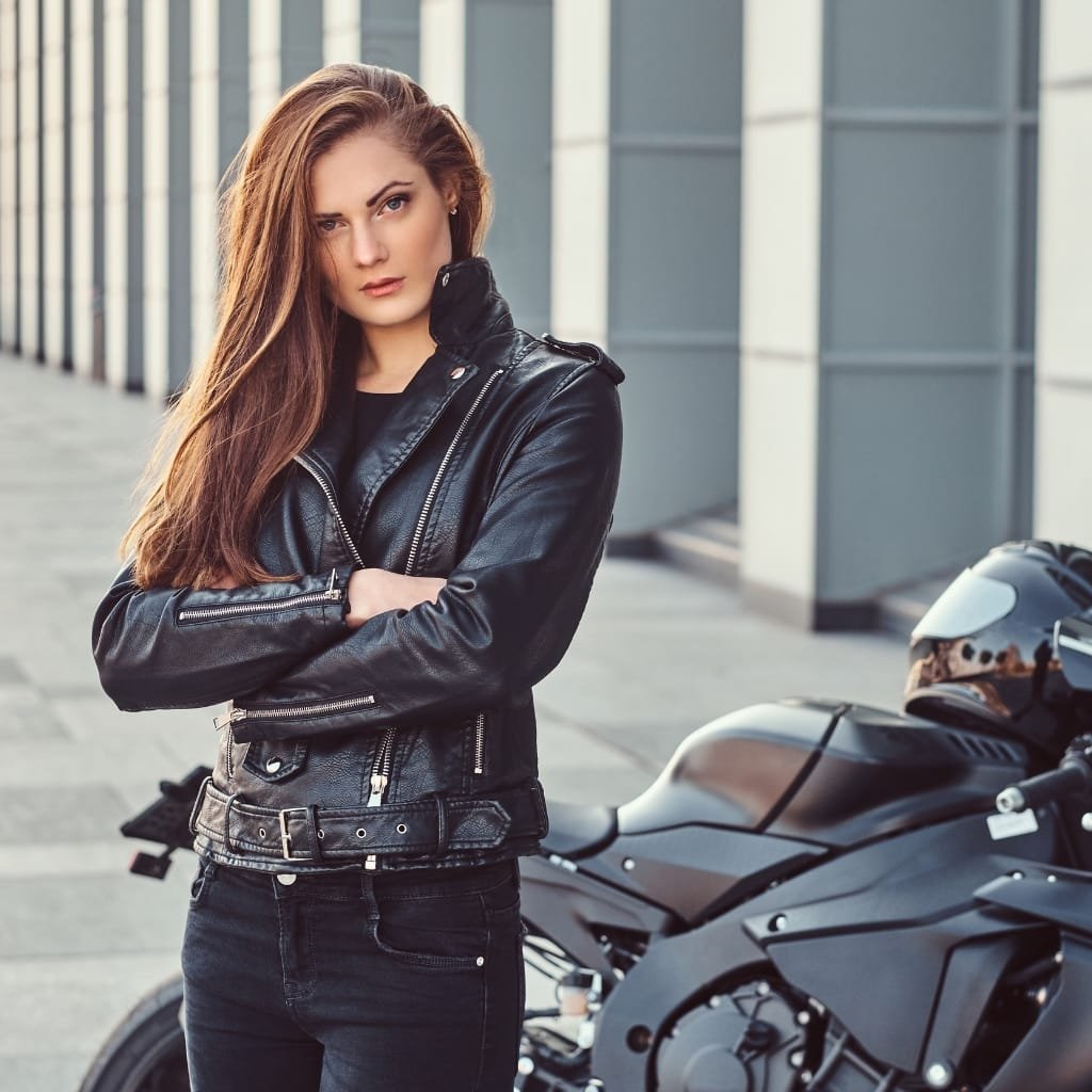 A woman with long, brown hair wearing a black Buffalo biker jacket, standing next to a motorcycle in an urban setting.