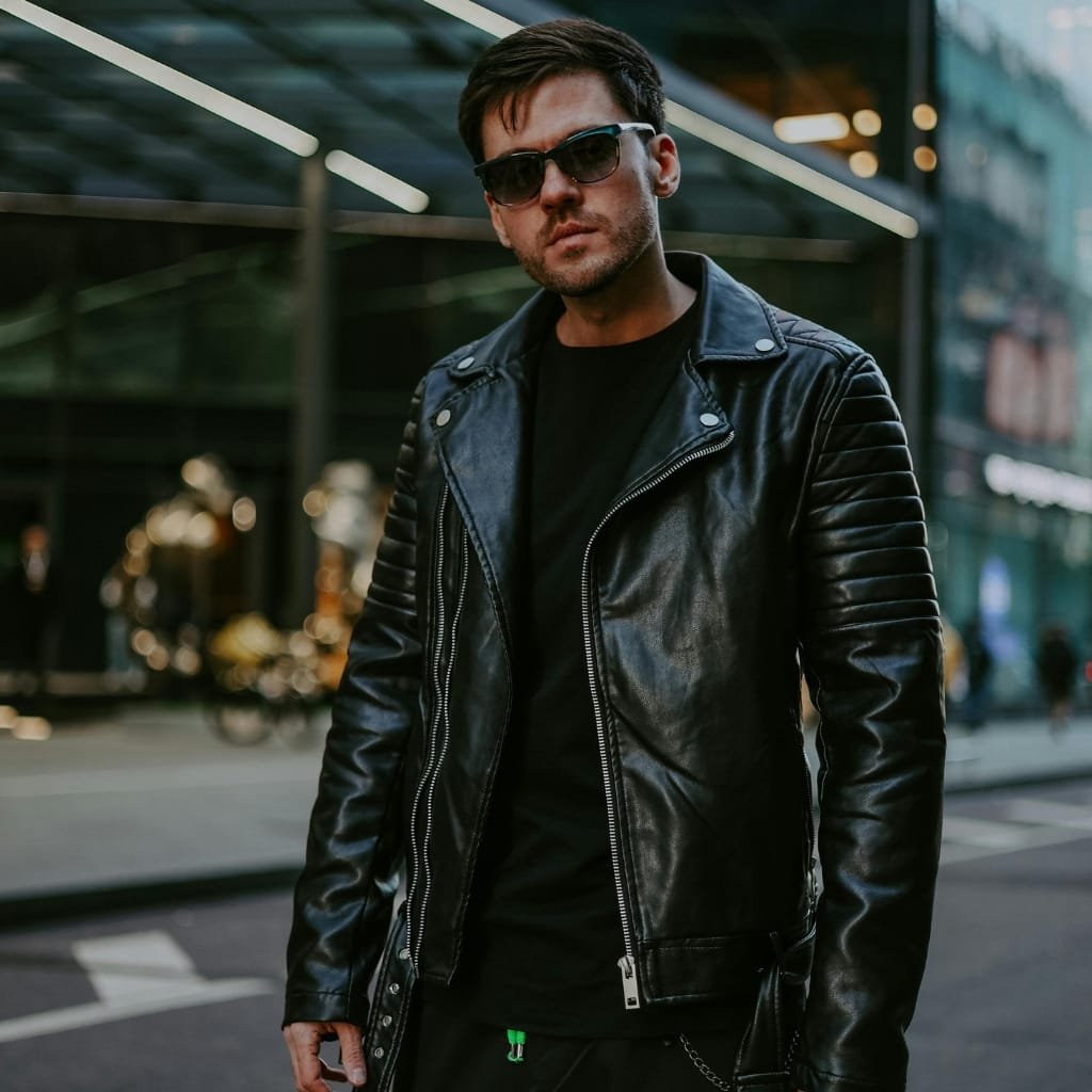 A young man standing outdoors in an urban setting, wearing a black A-1 bomber jacket with quilted shoulder details, black sunglasses, and casual black clothing underneath, exhibiting a street style fusion. The background features modern architecture and blurred city life.

