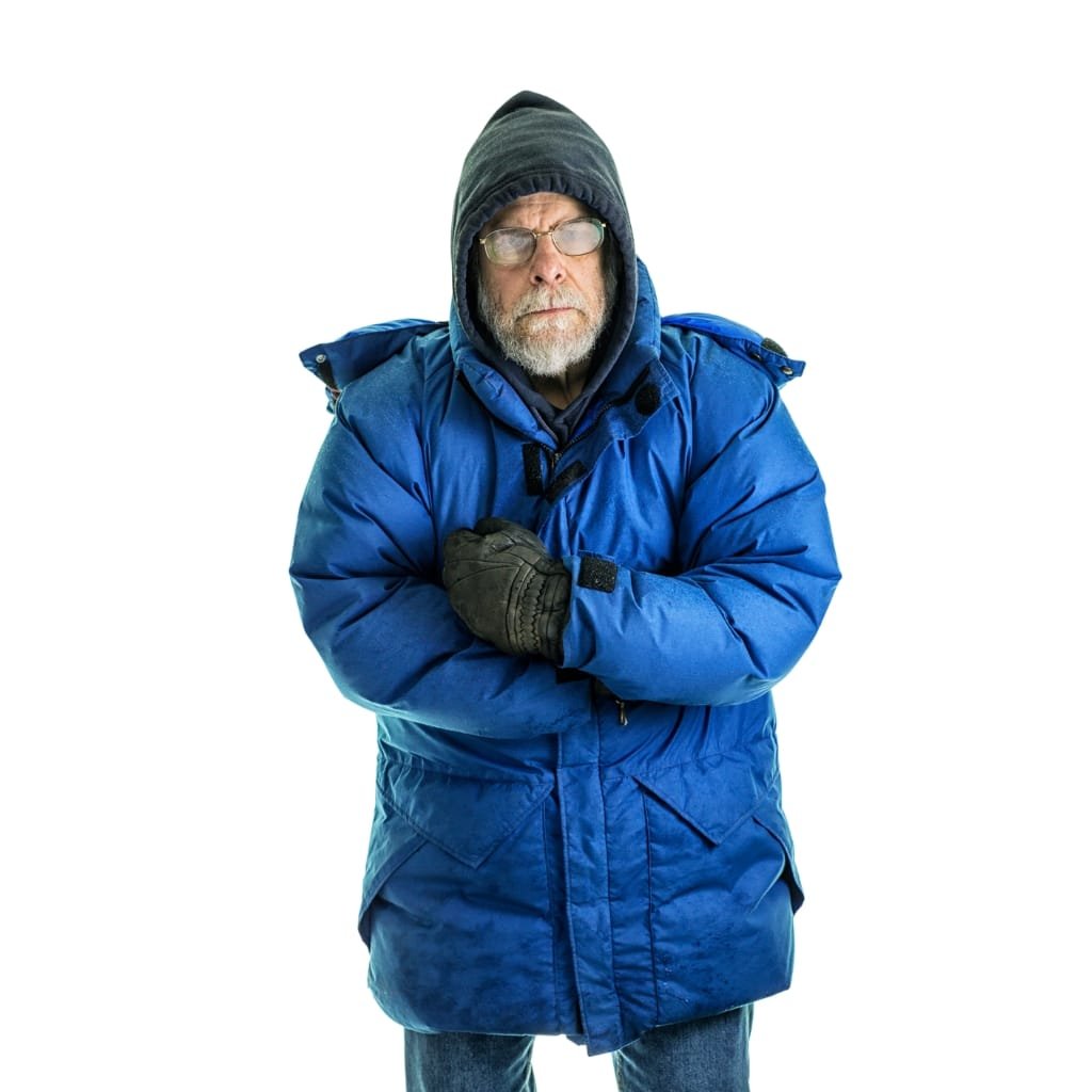 An older man dressed in a blue parka jacket, layered with a hoodie and gloves, standing against a plain white background, looking serious.

