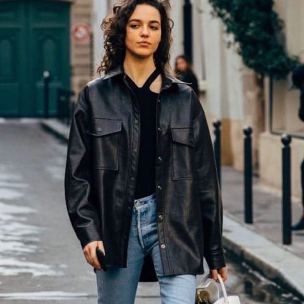 A woman walking on a city street wearing a black cowhide leather shirt jacket, paired with light blue jeans and holding a small bag.