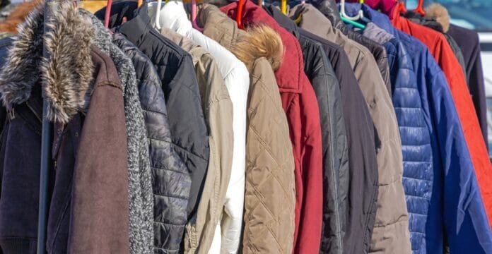 A row of various winter coats hanging on a clothing rack outdoors, including different styles and colors such as brown, black, white, red, and blue, with some featuring fur-lined hoods.