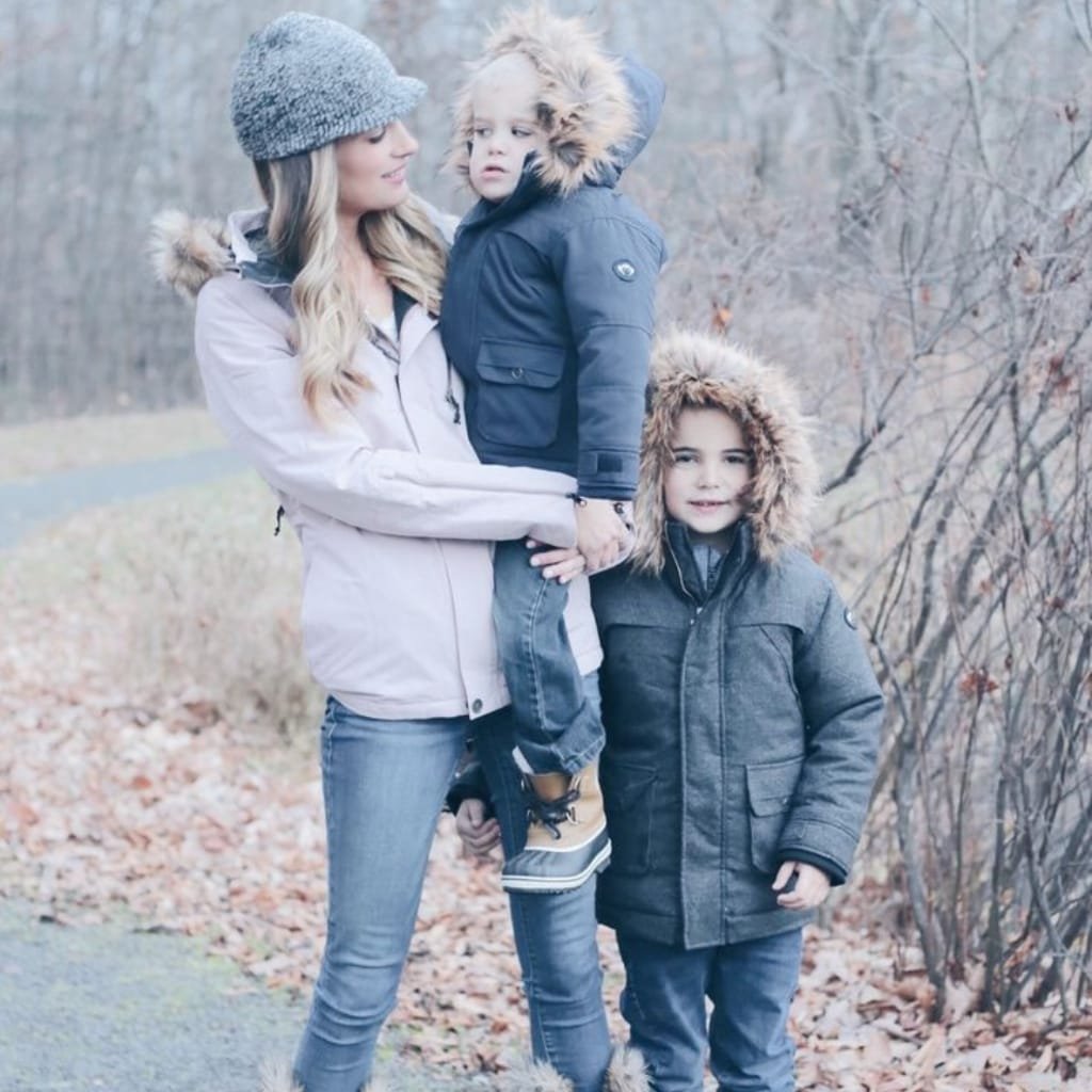 Alt Text:
A woman and two young boys dressed in warm winter coats and boots stand on a forest path during a chilly day. The coats and boots are fur-lined and designed for cold weather.

