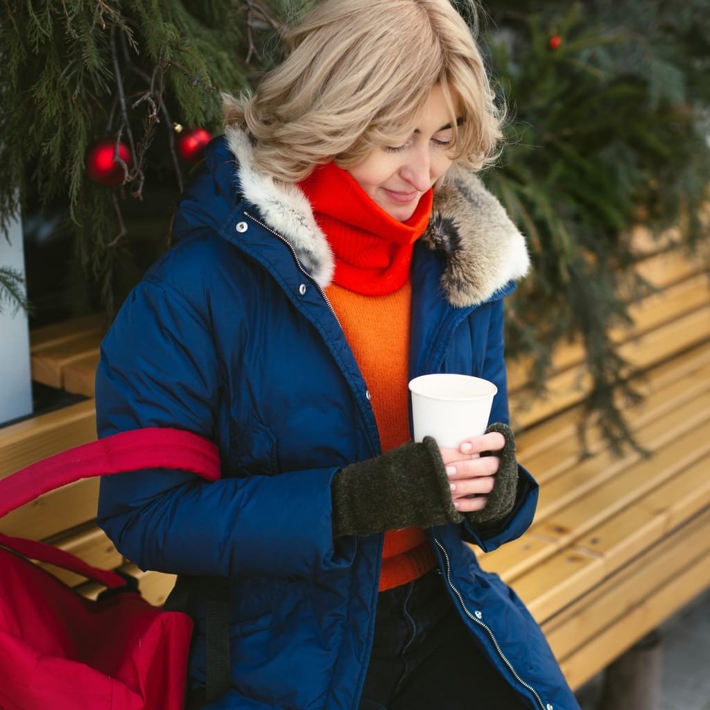 Woman in a blue Alaska winter coat sitting on a bench, holding a hot drink in a city park.