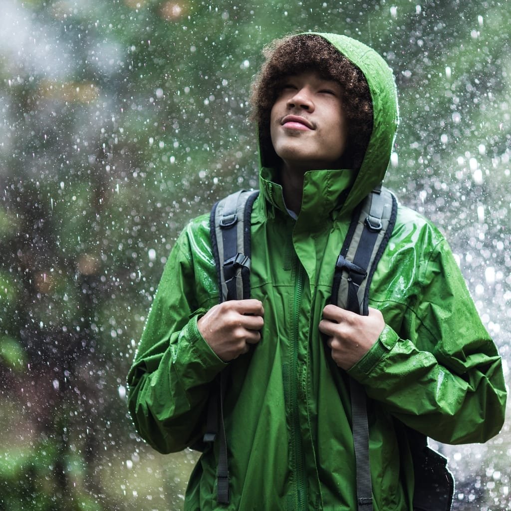 A person wearing a bright green anorak jacket with the hood up, walking outdoors in the rain with water droplets splashing around.

