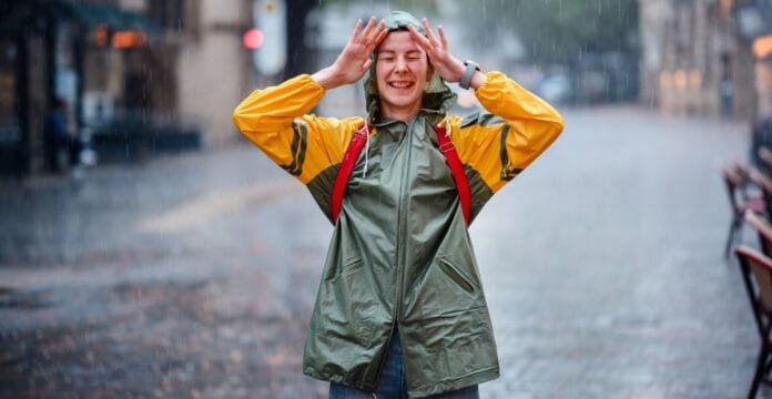 A woman wearing a green and yellow waterproof rain jacket, smiling and holding her hands to her head as rain falls around her on a city street.