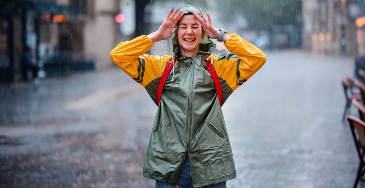 How to Style an Anorak Rain Jacket for Every Occasion A woman wearing a green and yellow waterproof rain jacket, smiling and holding her hands to her head as rain falls around her on a city street.