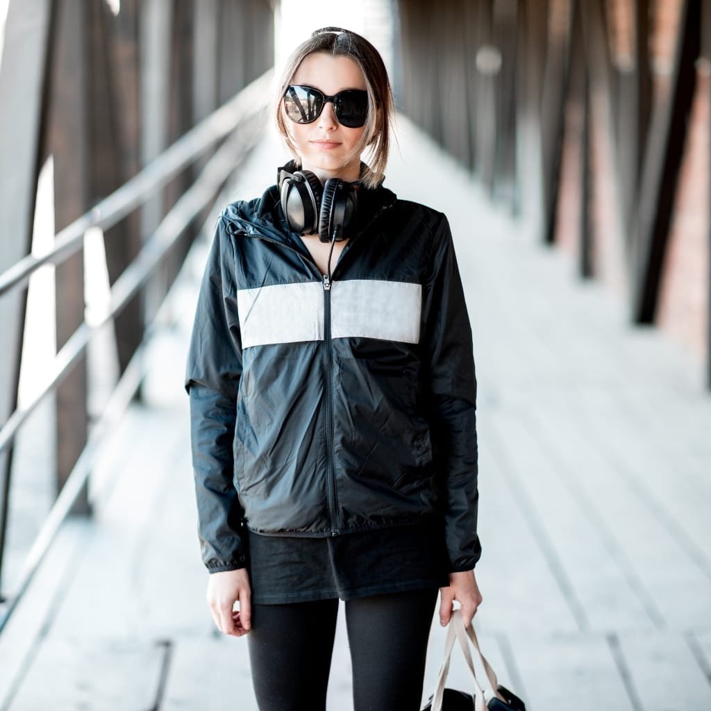 A woman standing outdoors on a bridge, wearing black athleisure wear with a black and gray rain jacket with a hood, black sunglasses, and headphones around her neck, holding a tote bag.