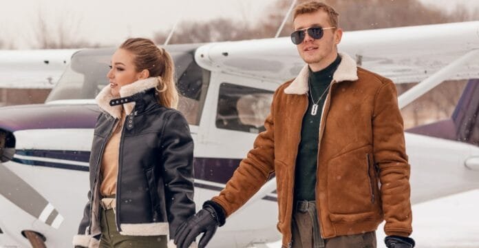 A man and a woman dressed in stylish aviator jackets standing beside a small airplane on a snowy tarmac, holding hands.
