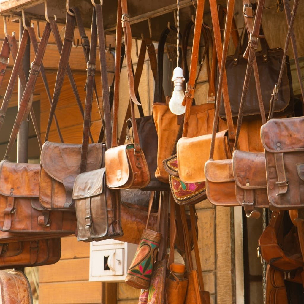 A display of various leather bags and wallets hanging on hooks in a rustic shop. The collection includes shoulder bags, crossbody bags, and small wallets in different shades of brown, tan, and black.

