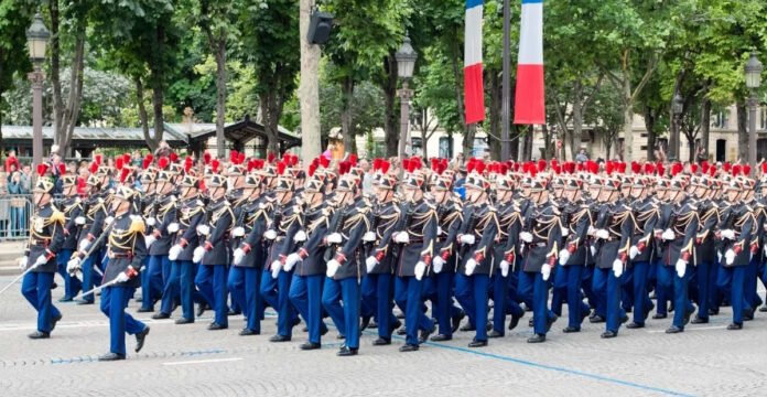 A large parade of uniformed soldiers marching in formation on a city street, decorated with trees and French flags. The soldiers are wearing dark jackets with gold accents, blue trousers, white gloves, and tall red plumes on their hats.