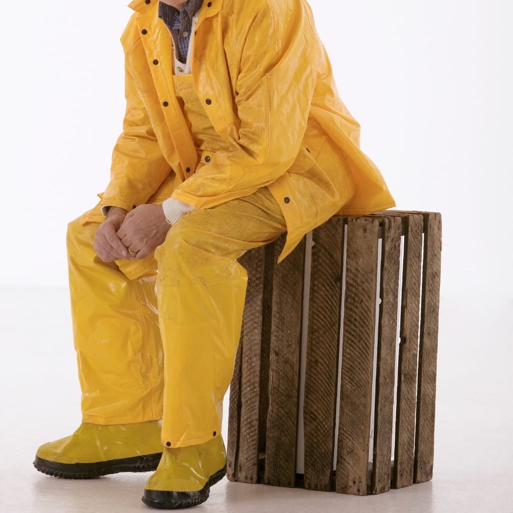 A person dressed in bright yellow rain gear, including waterproof pants, jacket, and boots, sitting on a wooden crate against a plain white background.

