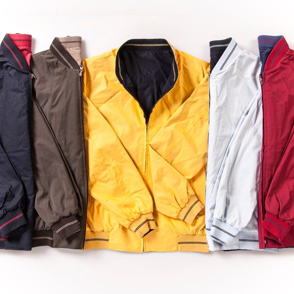 A row of five colorful minimalist bomber jackets laid flat on a white surface, featuring navy blue, brown, bright yellow, white, and red jackets.