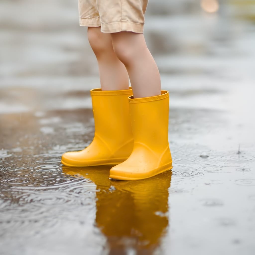 Child wearing bright yellow waterproof rain boots standing in shallow puddle on a rainy day.

