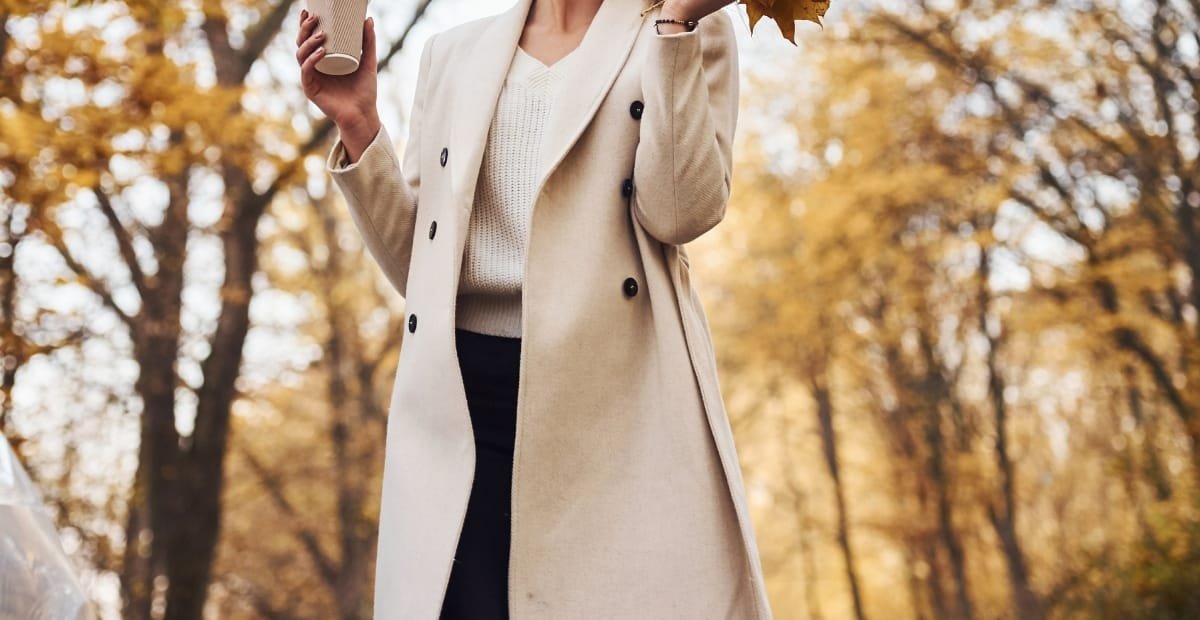 Best Ladies Wool Coats for Timeless Warmth and Style A woman standing outdoors in autumn, wearing a stylish beige wool coat over a white sweater, holding a coffee cup, surrounded by fall foliage.