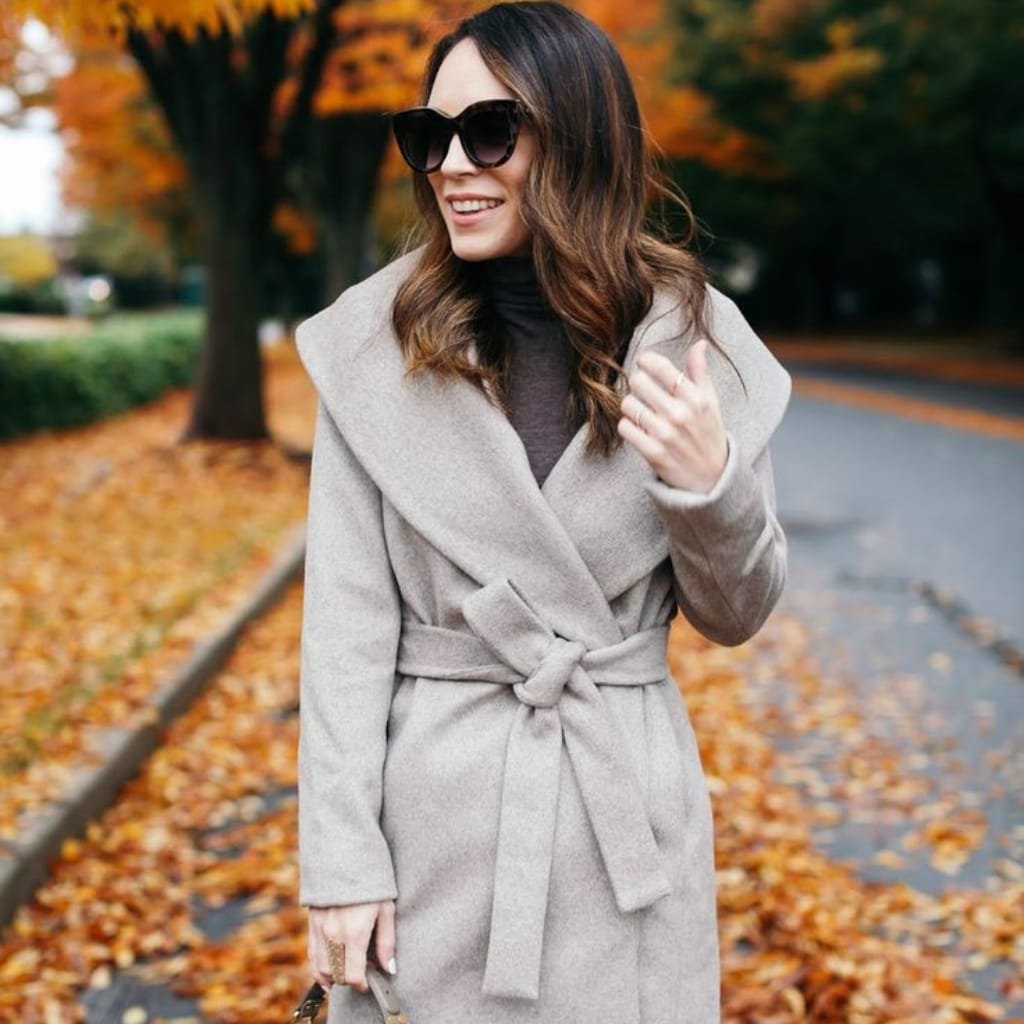 A stylish woman outdoors on a fall day wearing a light-colored wrap coat with a belt, black turtleneck, and large sunglasses, surrounded by autumn foliage and fallen leaves.