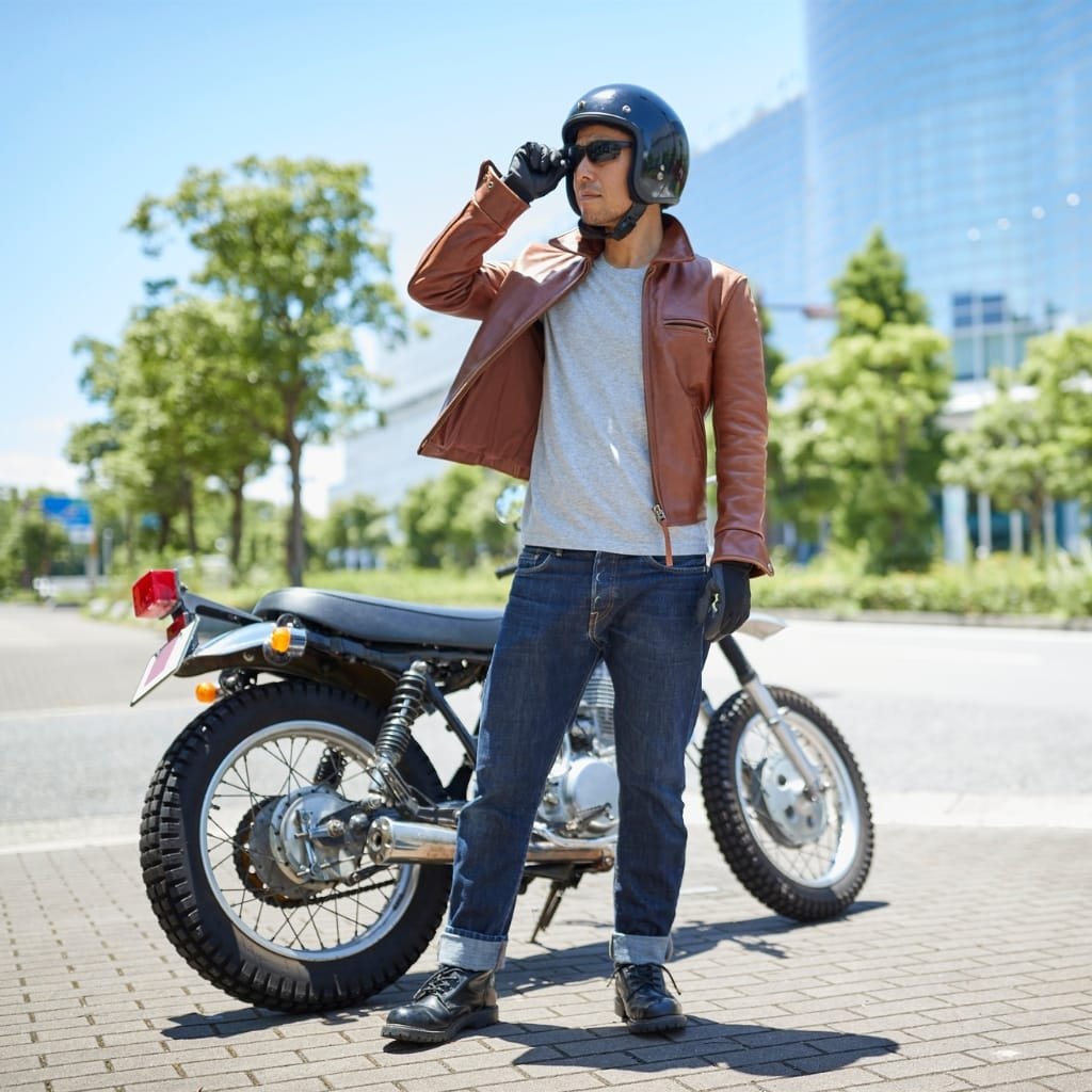 A man wearing a brown leather biker jacket, jeans, and black boots, standing next to a motorcycle outdoors on a sunny day.

