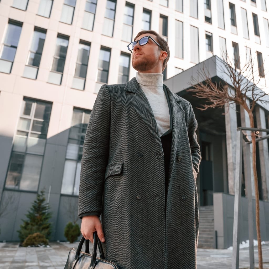 A stylish man wearing a gray wool coat with a herringbone pattern, dark sunglasses, a light turtleneck sweater, and carrying a black leather briefcase, standing outside in an urban setting with modern office buildings.