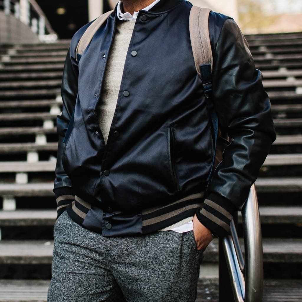 A person wearing a black Buffalo bomber jacket with striped cuffs and collar, standing on outdoor stairs with one hand in their pocket and a backpack over their shoulder.