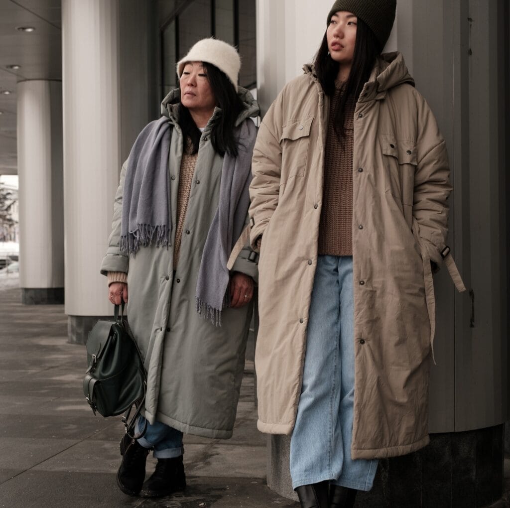 Two women dressed in long winter coats, standing outdoors near a building, both wearing oversized, calf-length coats in neutral tones, accessorized with hats and casual footwear.