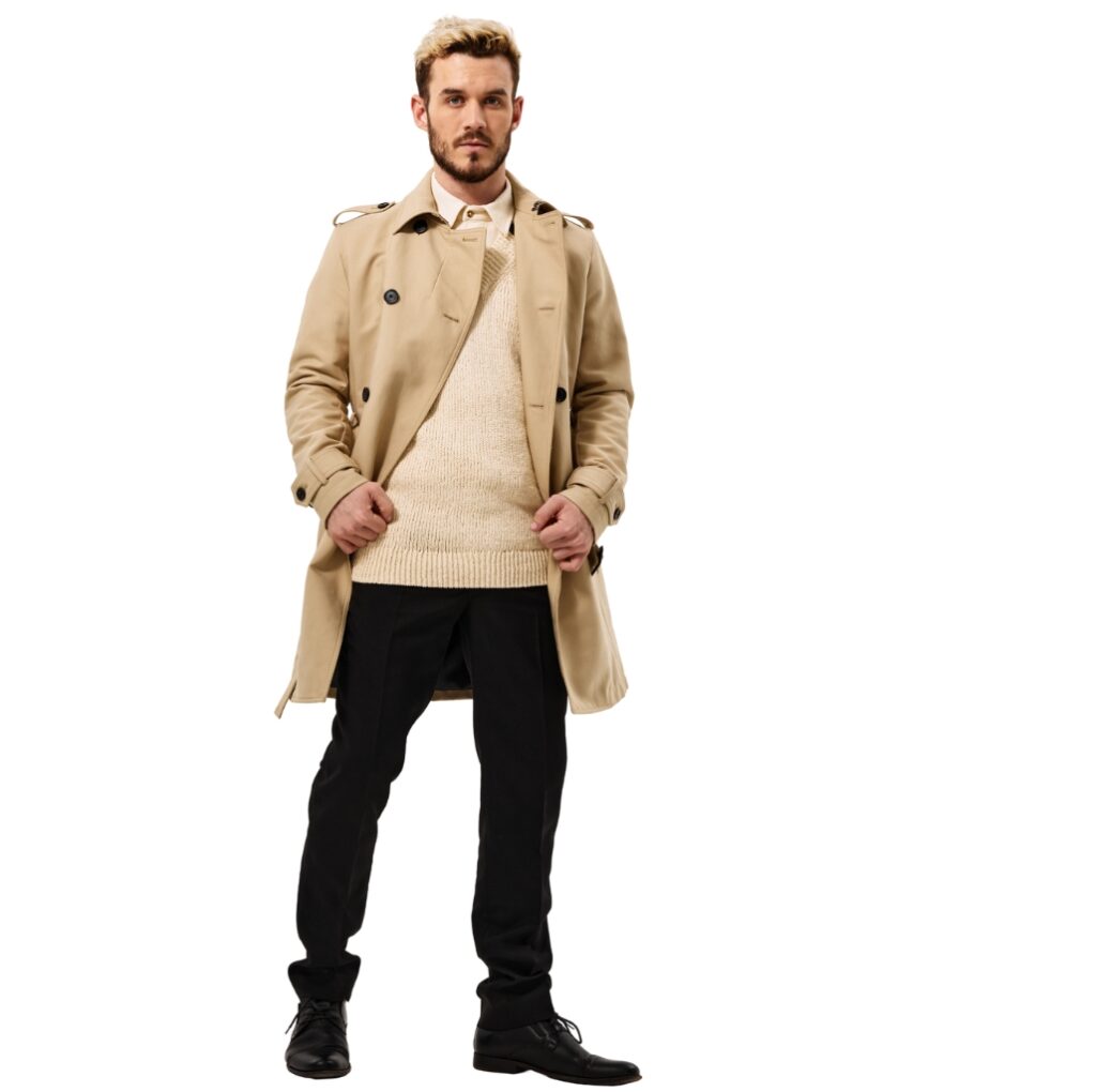 A man standing against a white background, wearing a beige calf-length winter coat, black pants, and black shoes. He has a beard and is holding his coat with both hands.