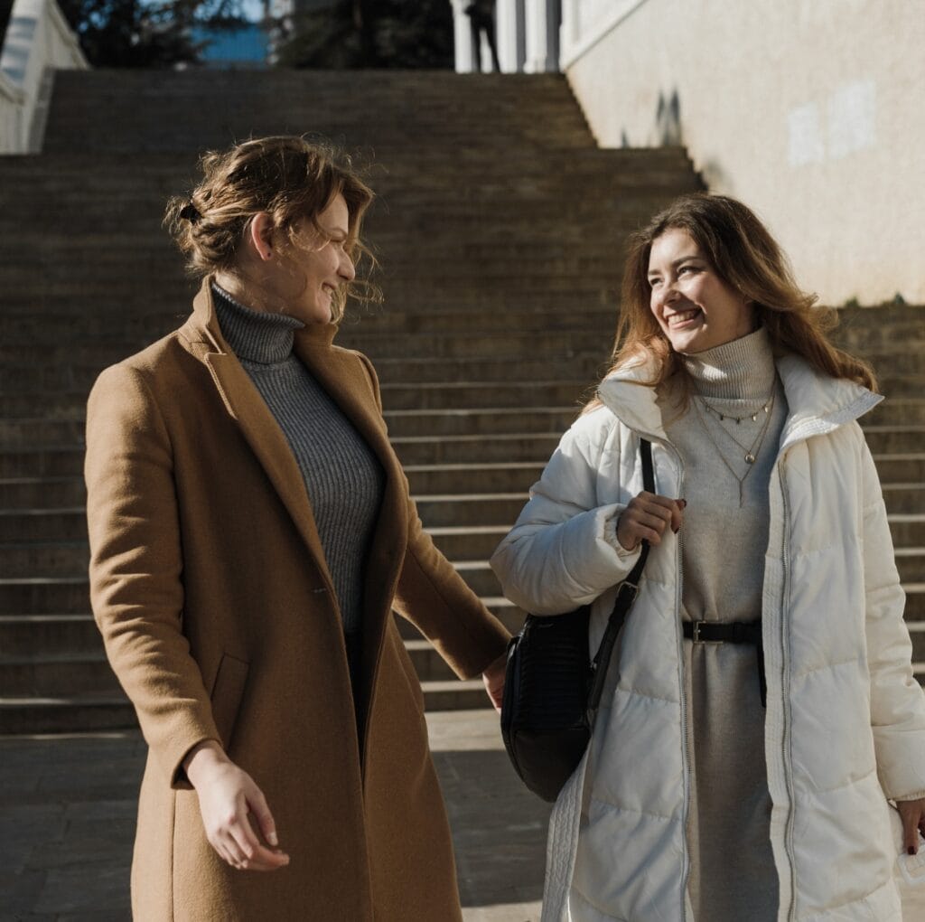 Two women walking outdoors on stairs; one wearing a long brown winter coat and the other a white puffer coat, both smiling and engaging in conversation.