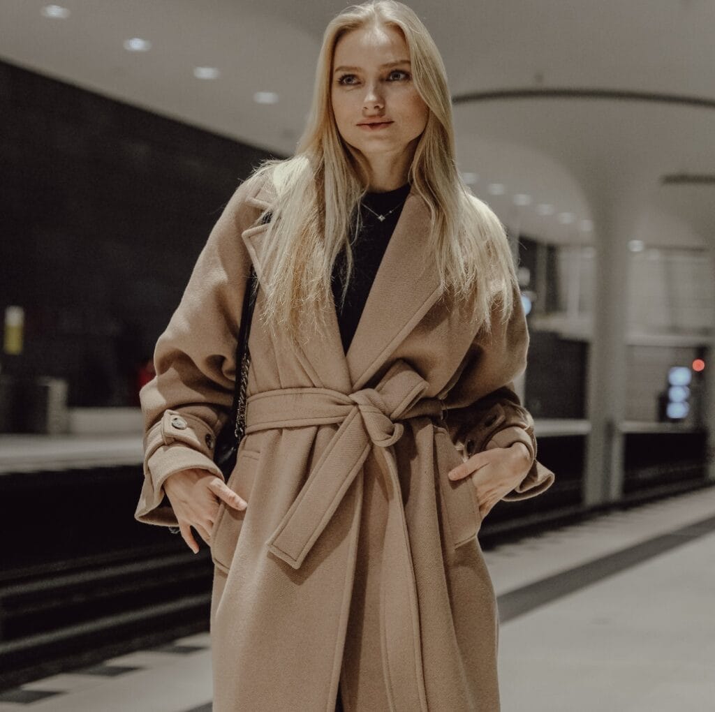 A woman wearing a tan calf-length winter coat with a belt, standing in a modern indoor setting.