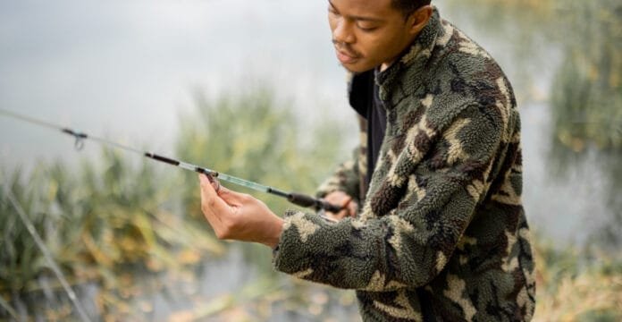 A person wearing a camouflage wool coat while fishing near a body of water with reeds and greenery.