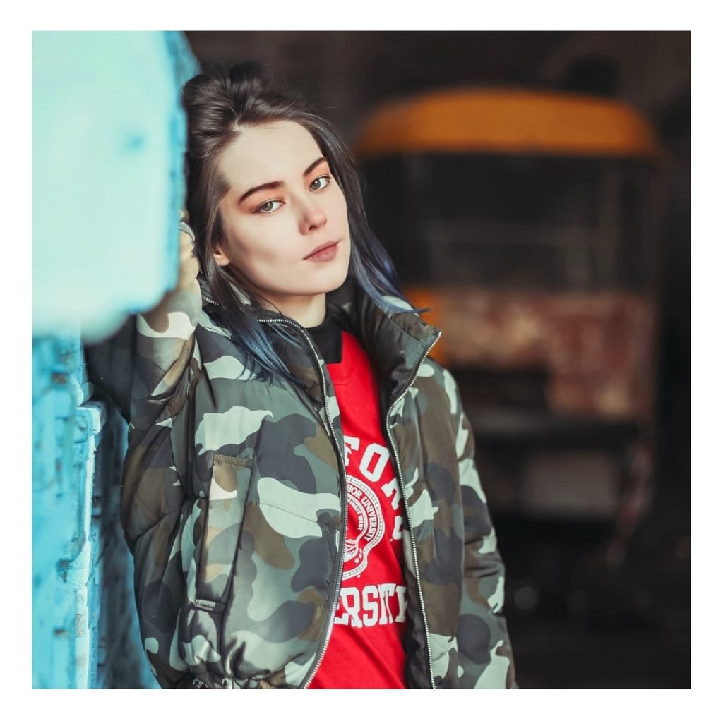 A young woman with dark hair and fair skin, wearing a camouflage bomber jacket, leaning against a blue wall with an urban background.