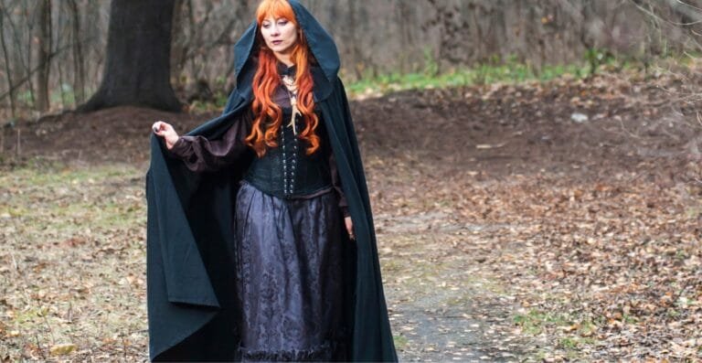 A woman dressed in a long, dark cape coat with a hood, standing in a wooded outdoor setting during autumn.