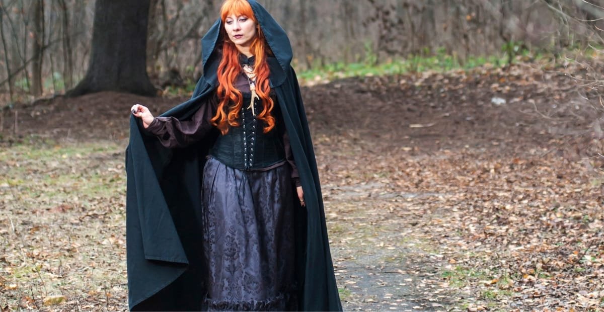 A woman dressed in a long, dark cape coat with a hood, standing in a wooded outdoor setting during autumn.