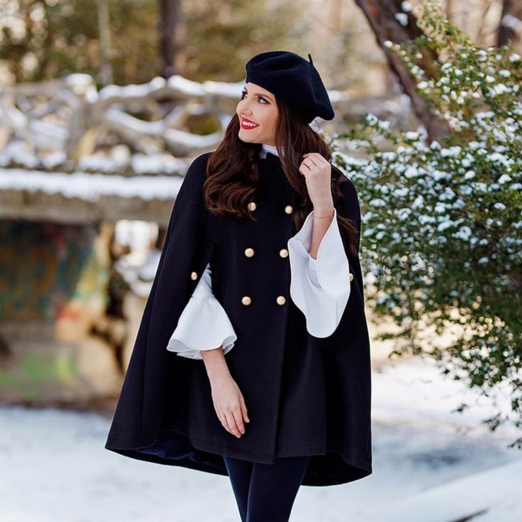 A woman standing outdoors in a winter setting, wearing a black cape coat with white bell sleeves, a black beret, and vibrant red lipstick, smiling and looking off to the side.