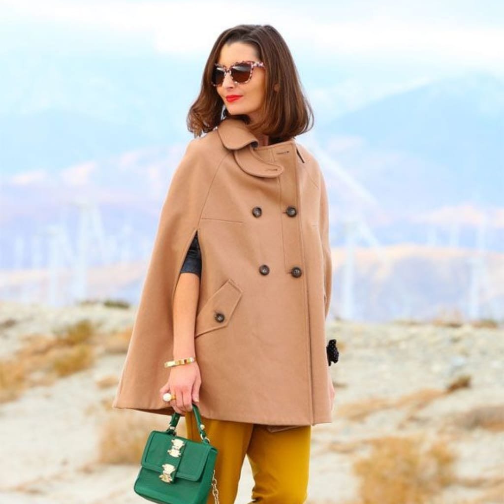A stylish woman standing outdoors in a scenic landscape, wearing a tan cape coat with button details, yellow pants, and sunglasses. She holds a small green handbag, accessorized with jewelry, and has a confident, fashionable pose.