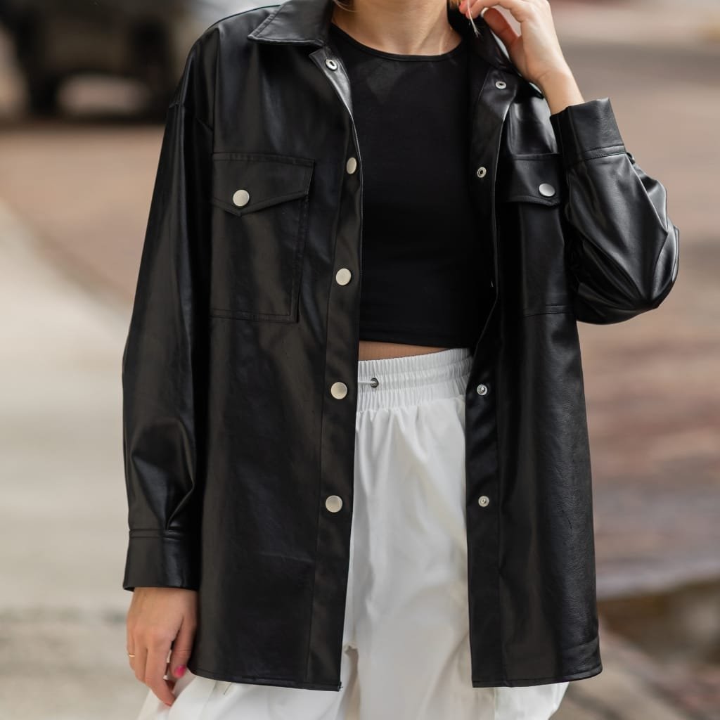 A person wearing a black faux leather oversized shirt or jacket over a black crop top, paired with white drawstring pants, standing outdoors on a casual day. The person has their left hand near their face and is holding a strand of hair, with a blurred background.

