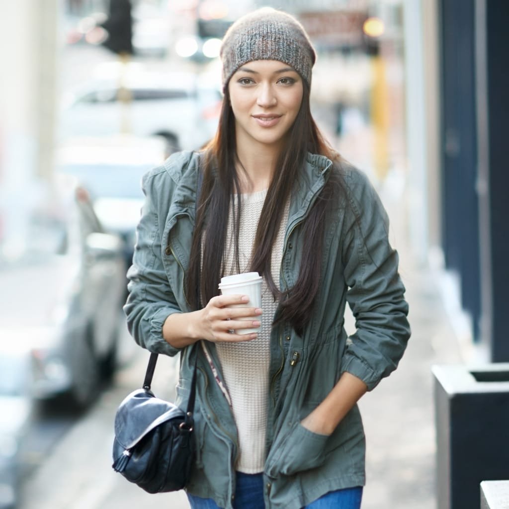 A young woman walking outdoors on a city street, wearing a casual gray jacket, a knit beanie, and holding a coffee cup, with her hand in her pocket.
