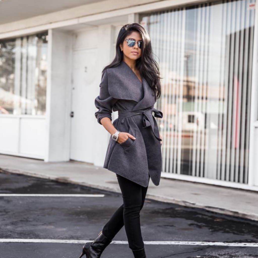 A stylish woman walking outdoors in casual attire, wearing a dark grey wrap coat, black skinny jeans, black high-heeled boots, and sunglasses with a cityscape background.