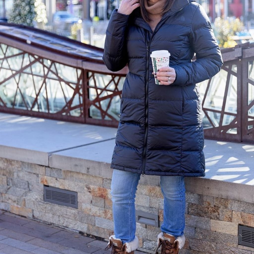 A person standing outdoors in an urban environment wearing a dark navy, puffy winter coat, holding a takeaway coffee cup. The person is standing next to a stone wall with a decorative metal railing, dressed in casual jeans and furry winter boots.

