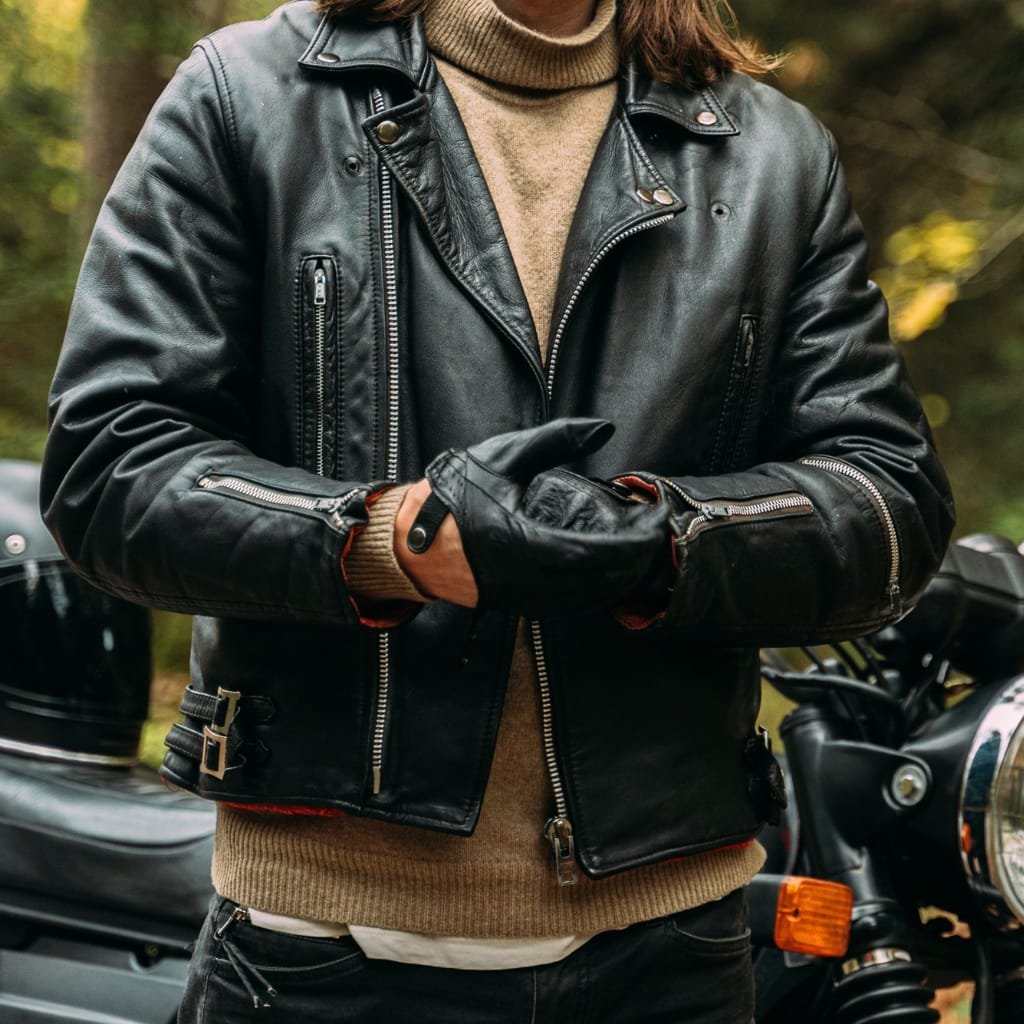 Top Racer Jacket Looks That Never Go Out of Style 6 A person wearing a classic black leather racer jacket, layered over a beige sweater and standing outdoors near a motorcycle.