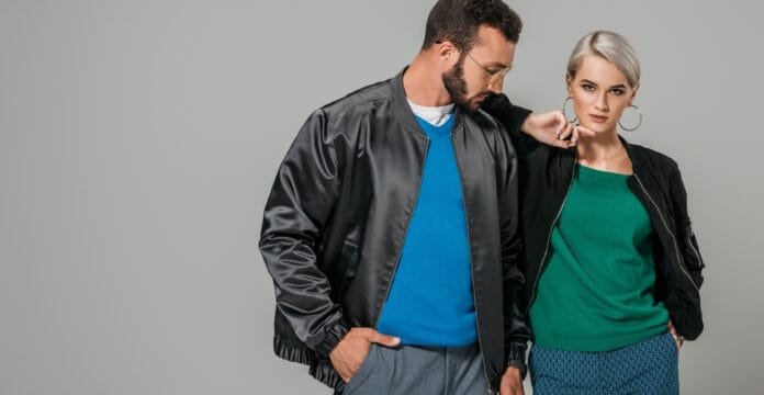A man wearing a classic black bomber jacket with a blue shirt underneath, standing beside a woman in a casual outfit, posing against a plain gray background.