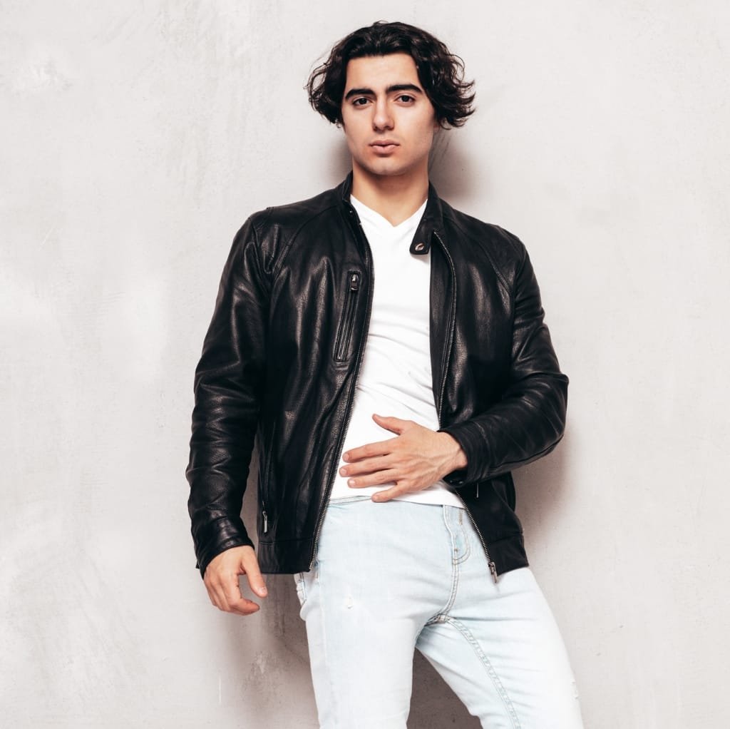 A person wearing a black leather bomber jacket over a white t-shirt, standing against a neutral background.
