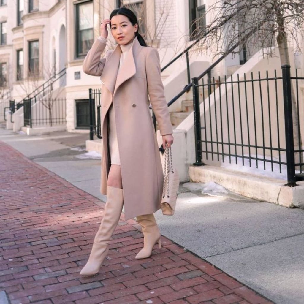 A woman standing outdoors on a sidewalk during winter, wearing a beige wrap coat over a cream dress, paired with thigh-high beige boots and carrying a beige handbag, with a cityscape background.