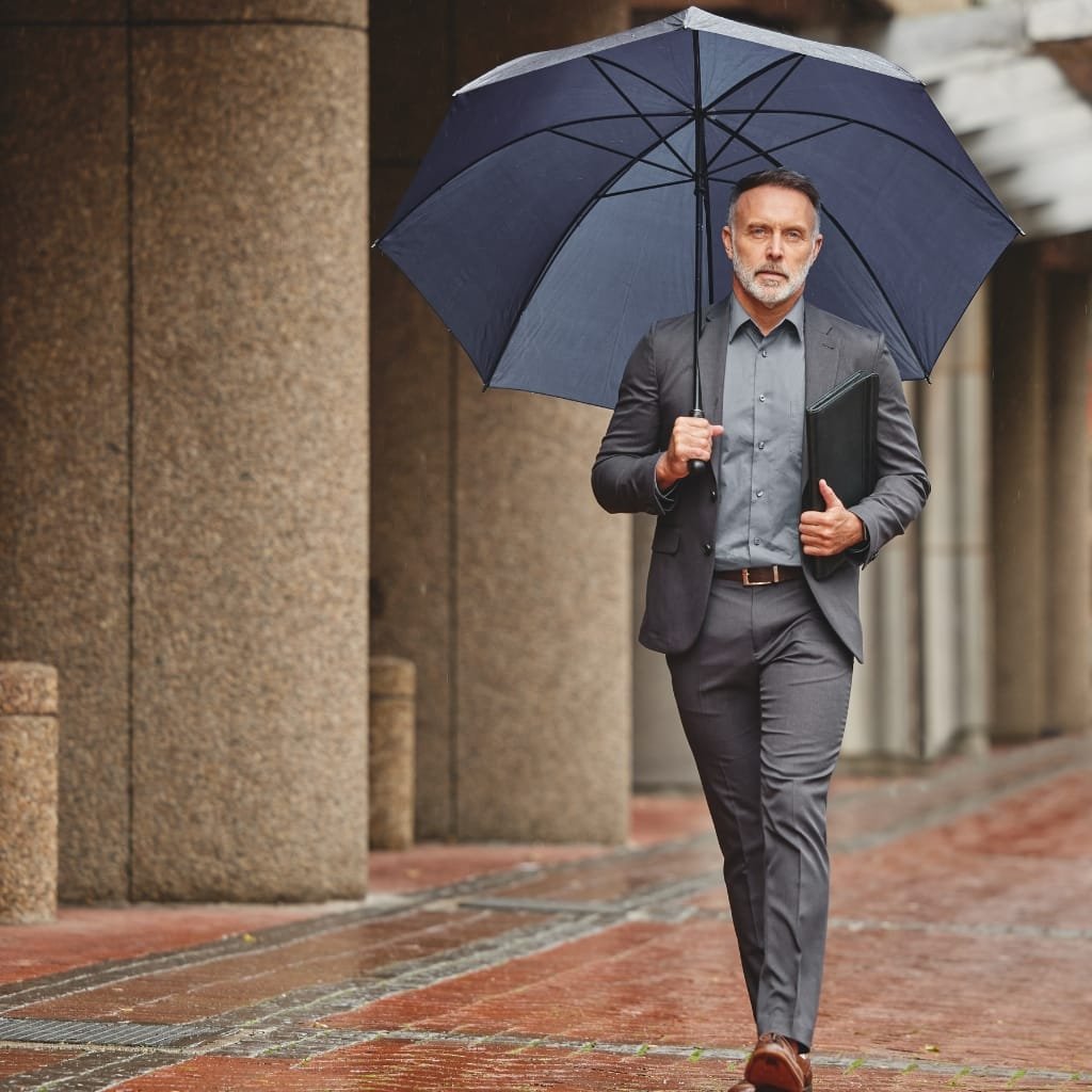 How to Style an Anorak Rain Jacket for Every Occasion 7 A man dressed in a stylish, dark grey suit holding a black umbrella, walking on a rainy city sidewalk while clutching a black folder.