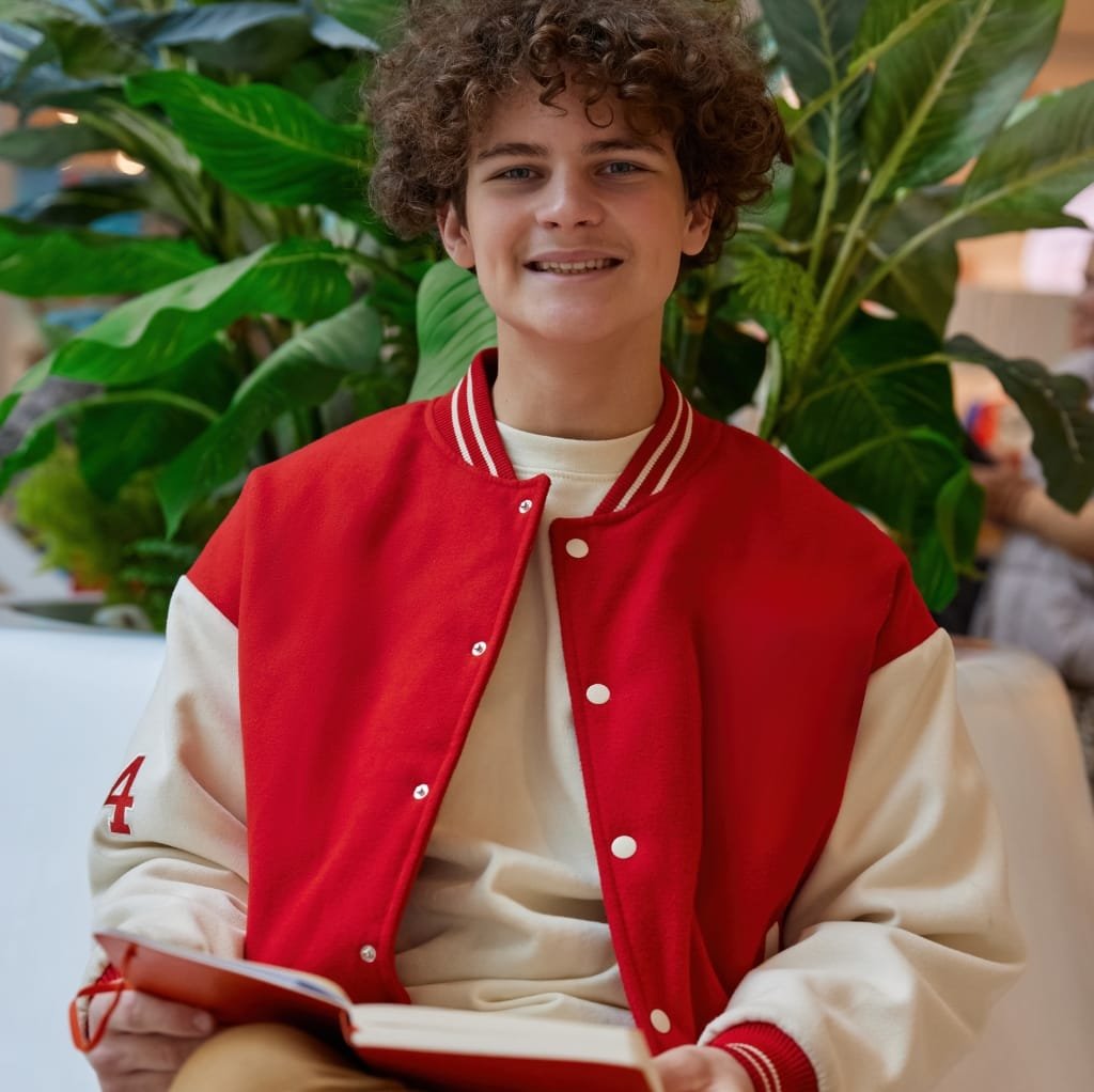 A young man with curly hair wearing a red and cream varsity jacket, sitting in front of large green plants, smiling at the camera.
