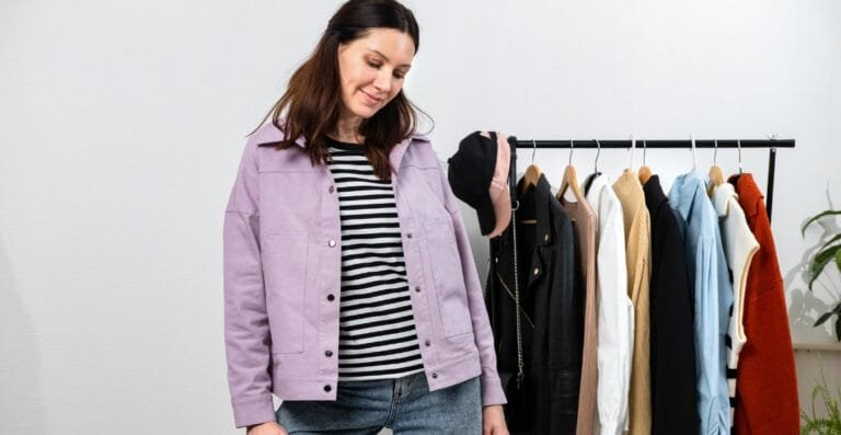 A woman looking at a rack of various clothing items while wearing a light purple cotton work jacket over a black and white striped shirt.