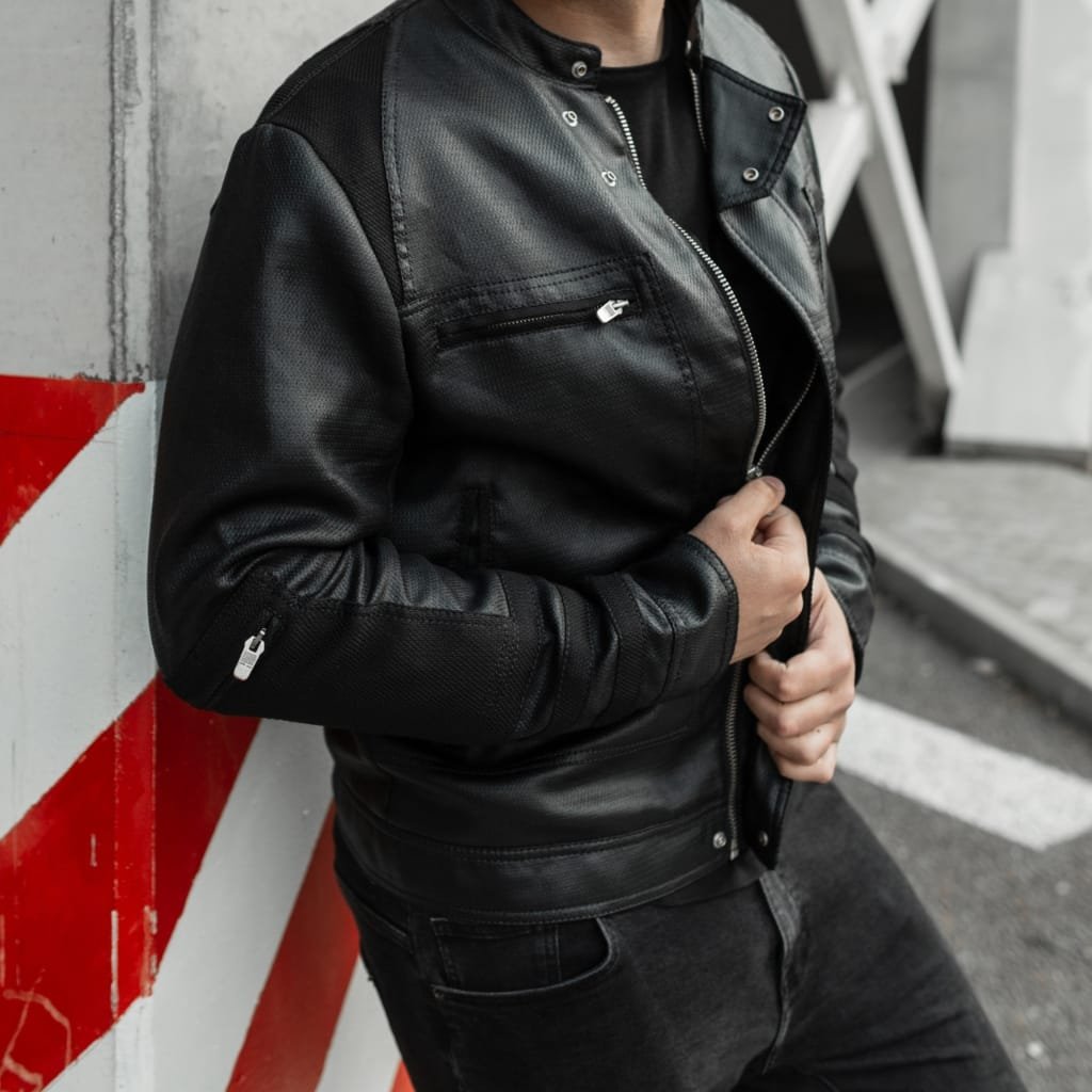 A person wearing a sleek black cowhide leather shirt jacket, standing against a concrete wall with red and white striped markings, with their hands adjusting the jacket's zipper.