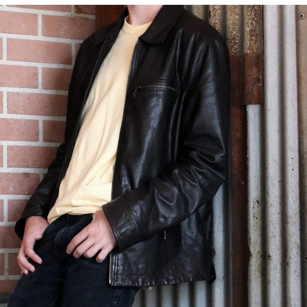 A person wearing a black cowhide leather shirt jacket, paired with a light-colored t-shirt and dark pants, standing against a brick wall and a corrugated metal background.

