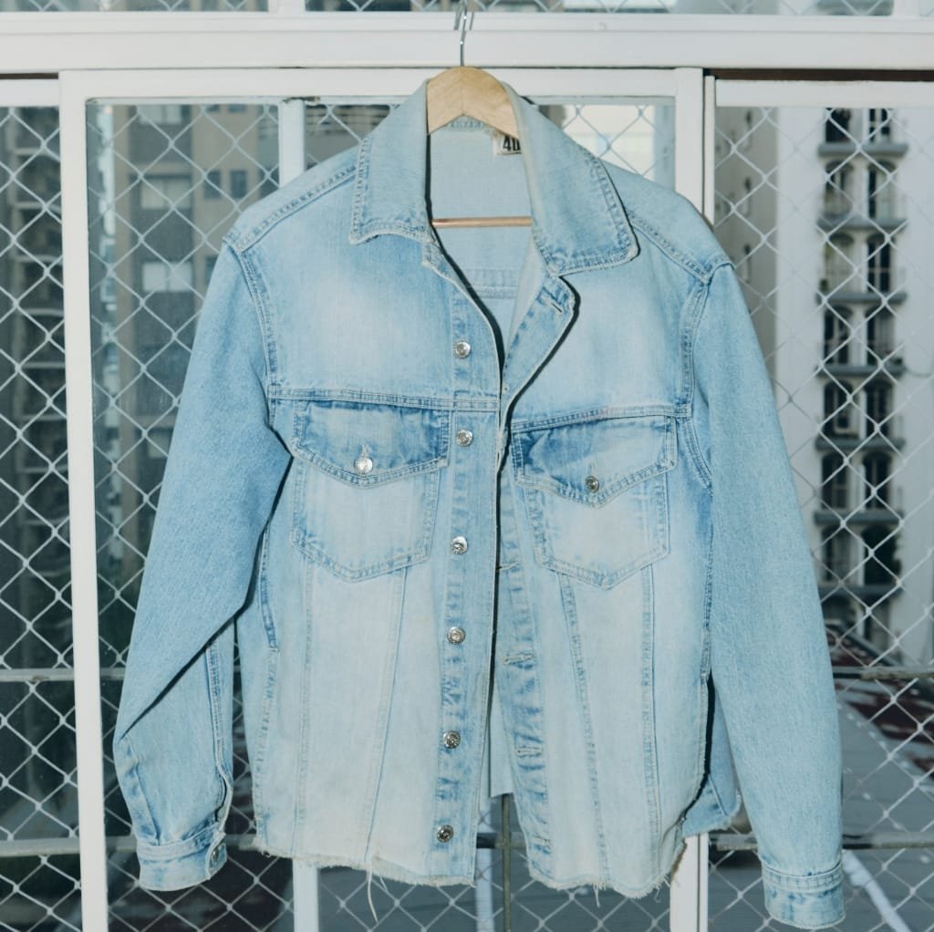 A light-wash denim bomber jacket hanging on a wooden hanger against a balcony railing with a cityscape in the background.
